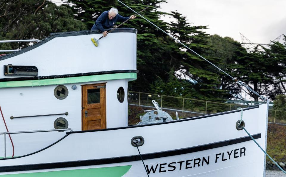 A man with white hair holds a long-handled tool a he cleans an exterior surface of a boat.