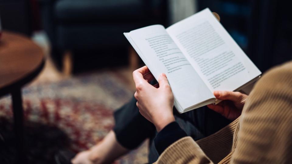 cropped shot of young asian woman taking a break from technology, relaxing and reading book on the sofa at a cozy home in the evening