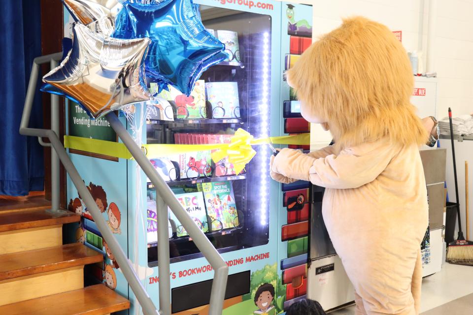 Merrill Elementary School in Raynham unveils its new Inchy the Bookworm vending machine, with a little help from Merrill the Lion, the school's mascot, who cut the bright yellow ribbon Thursday morning, May 27, 2022.