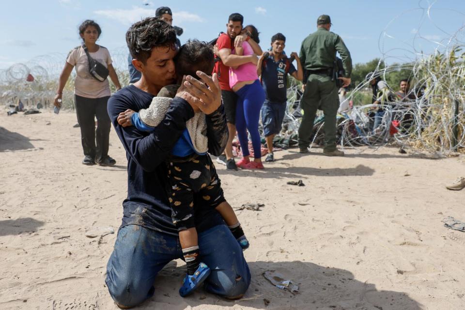 A man kneels on dry ground embracing a toddler as people climb through coils of wire behind him.