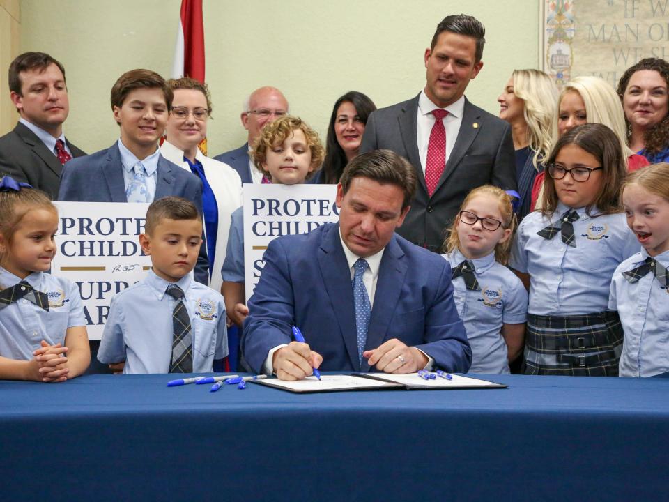 Ron DeSantis sitting at a table while he signs the so-called "Don't Say Gay" bill into law surrounded by young school children.
