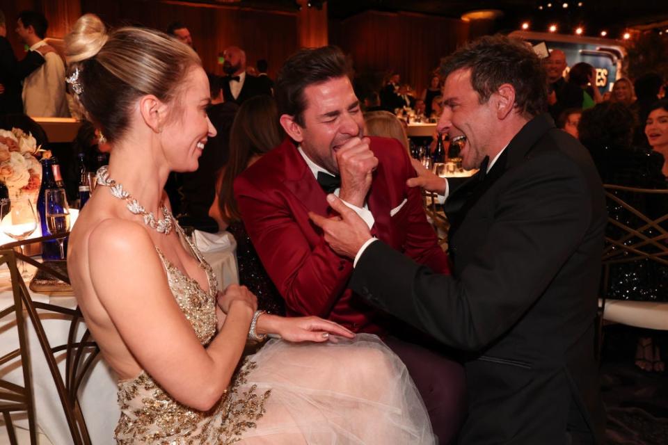 Three people engaged in a lively conversation at an event, one in a tuxedo, another in an elegant gown, laughing