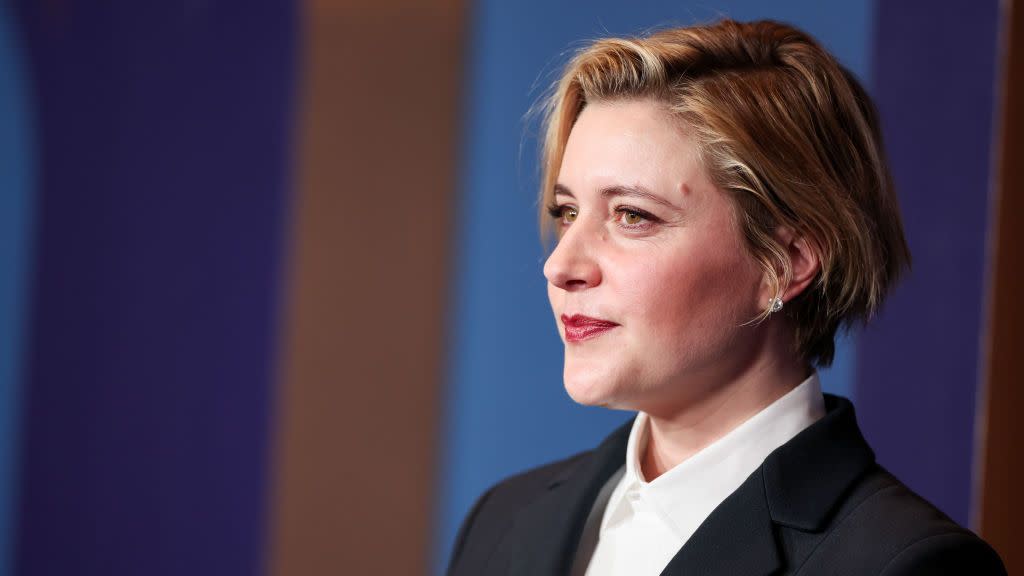 greta gerwig seen in profile looks out in front of her, she wears a white collared shirt and black suit jacket with stud earrings and makeup