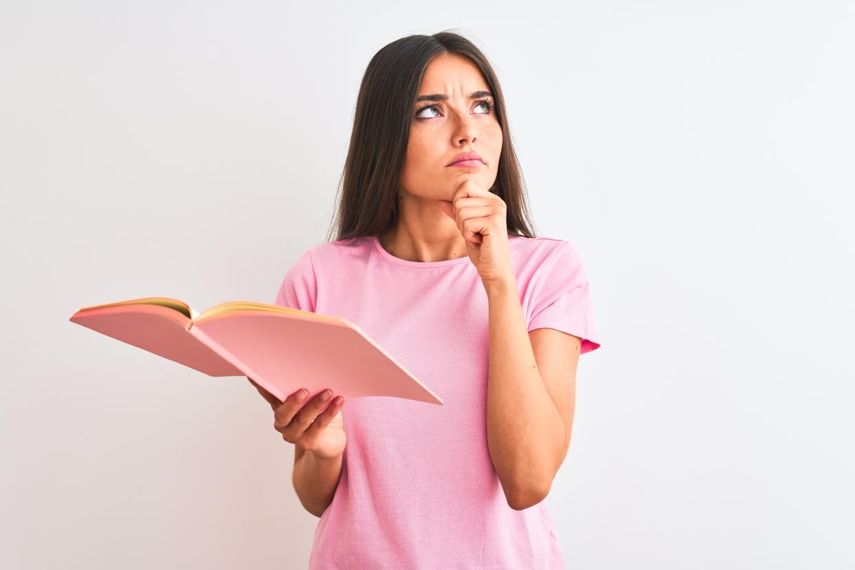 Young beautiful student woman reading book standing over isolated white background serious face thinking about question, very confused idea