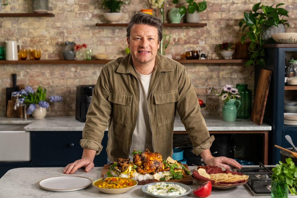 Dish - Kerala-style Roast.Chicken. Pictured: Jamie Oliver stood behind the counter with roast chicken, poppadoms and other plates of food surrounding the platter.