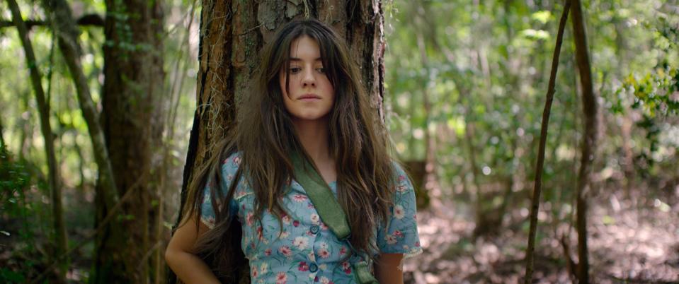 Woman standing in the woods, looking forward, with a tree trunk behind her