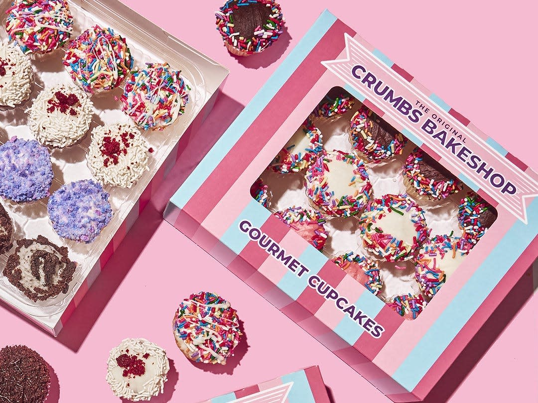 Boxes of cupcakes from the recently relaunched Crumbs Bake Shop appear against a pink background as a hand with red nails reaches for one from the lower right of the photo.