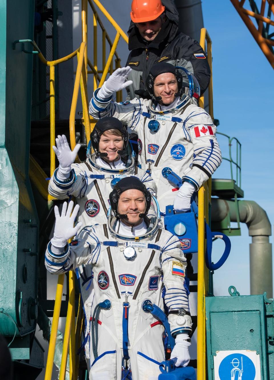 The Expedition 58 crew bids farewell to Earth as they wave to well-wishers at the base of their Soyuz rocket. From top, the crew is: Flight Engineer David Saint-Jacques of the Canadian Space Agency (CSA), top, Flight Engineer Anne McClain of NASA, center, and Soyuz Commander Oleg Kononenko of Roscosmos. <cite>NASA/Aubrey Gemignani</cite>