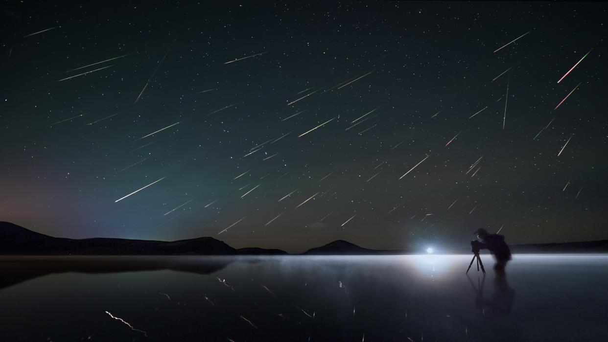  a person stands near a lake under a sky full of perseid meteors.  
