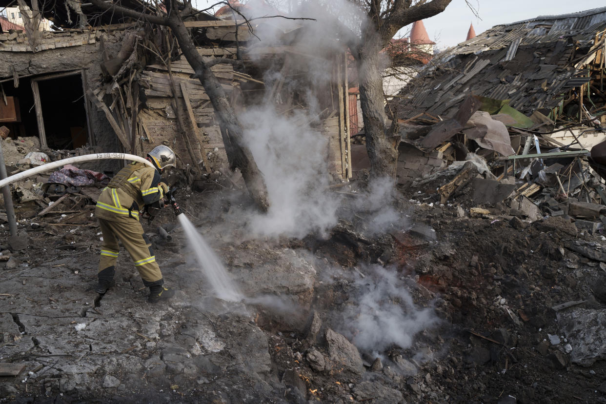 A firefighter trains a powerful hose on a fire next to houses destroyed in a Russian attack.