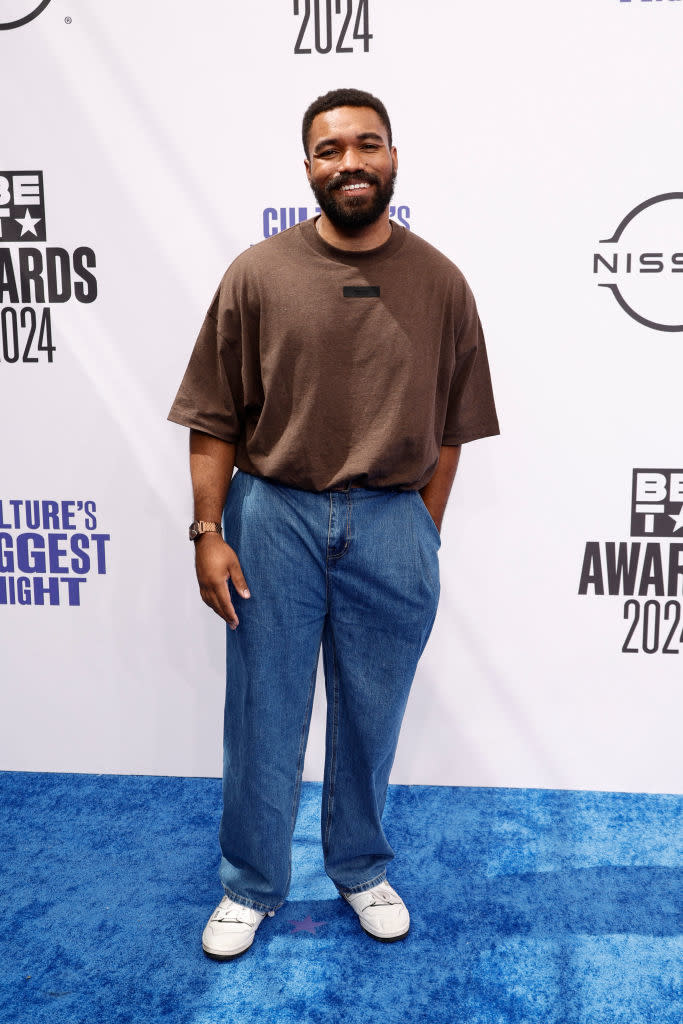 Jordan Howlett poses in casual attire with a brown t-shirt, loose blue jeans, and white sneakers on the blue carpet at the BET Awards 2024