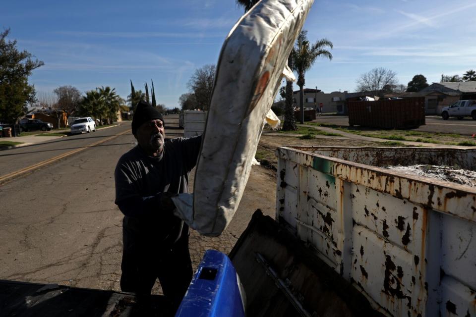 A man stands next to a large metal trash bin; a mattress is seen as it's tossed in the air.