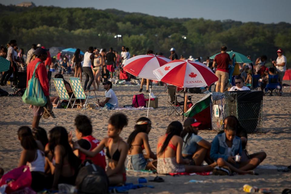 People gather on Toronto’s Woodbine Beach to celebrate Canada Day on July 1, 2020. In place of fireworks the federal government hosted an augmented reality experience that could be seen using a smartphone. See crowd at the beach, Canada beach umbrellas and a group of young women in fg. 
