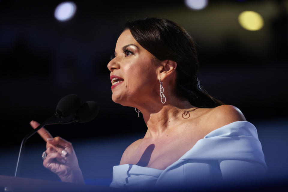 Ana Navarro speaking at a podium, wearing an off-shoulder dress and dangling earrings