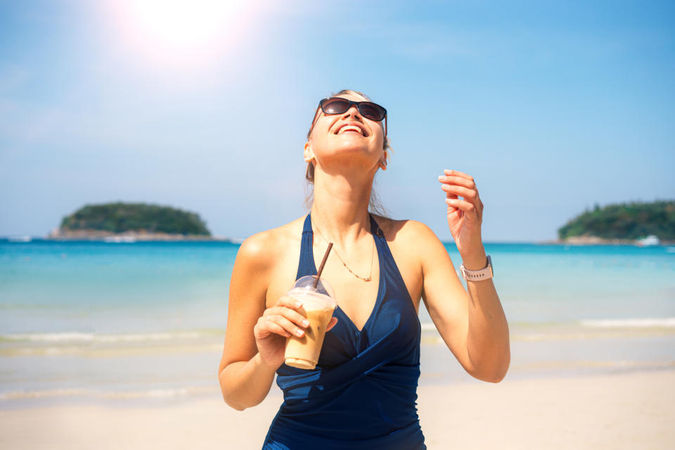 An image of a woman standing on a sunny beach wearing a navy v-necked swimsuit and tipping her head back to feel the sun on her face. She's wearing sunglasses