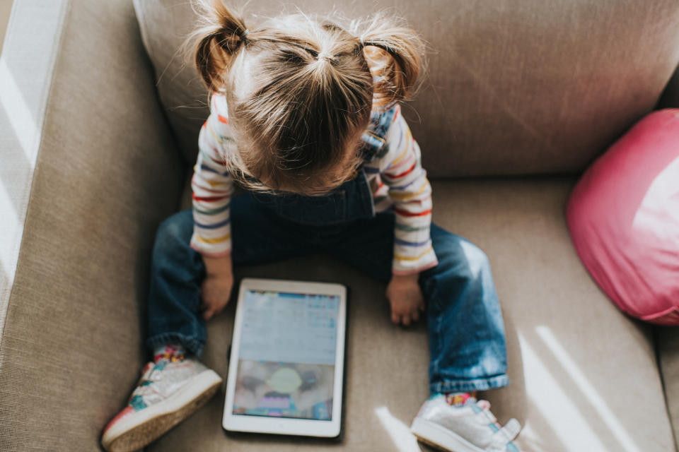 Child seated on sofa, looking at a tablet screen, indicative of technology's role in modern parenting