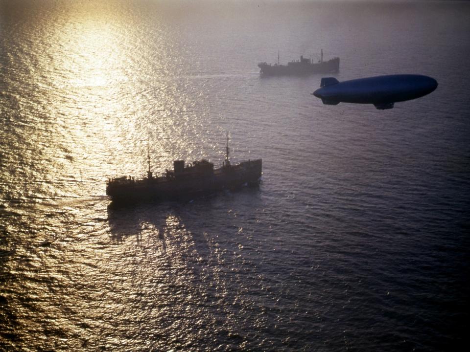 Two unidentified merchant cargo ships from a convoy are on the water below as they are escorted by a K-Class patrol blimp floating above as photographed in the 1940s.