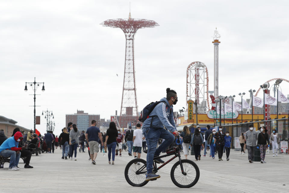 Un joven con cubrebocas pasea en bicicleta el domingo 24 de mayo de 2020 en el abarrotado paseo de Coney Island, durante la pandemia del coronavirus, en Nueva York. (AP Foto/Kathy Willens)