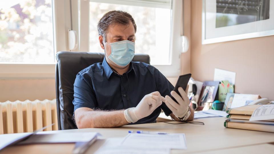 boss answering phone call with mask and gloves in office with protective mask and gloves holding mobile phone.