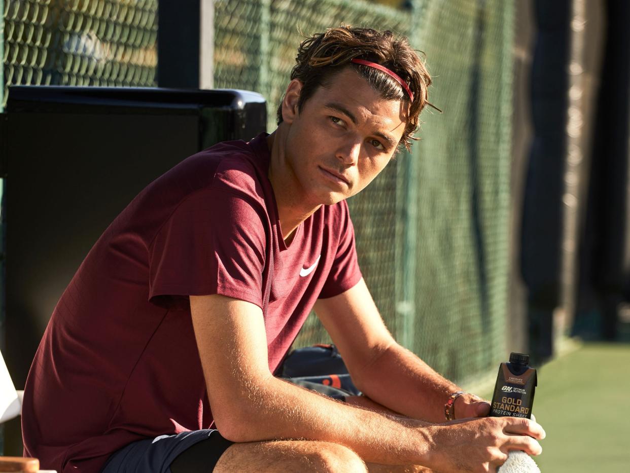 Tennis star Taylor Fritz sitting courtside in athletic clothes, holding a protein shake, with his tennis racket leaning on the bench next to him
