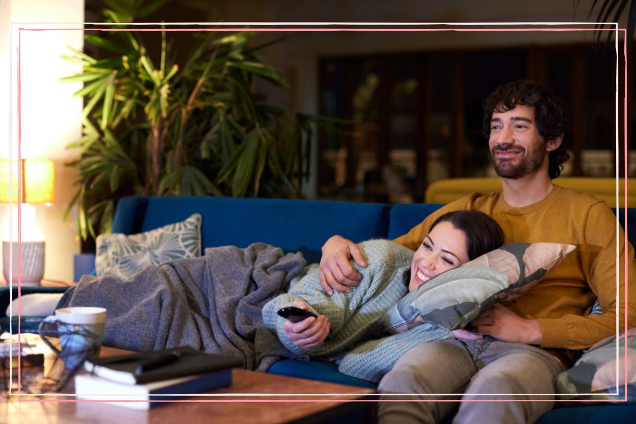 A couple cuddling on a sofa and pointing a remote at a TV. 