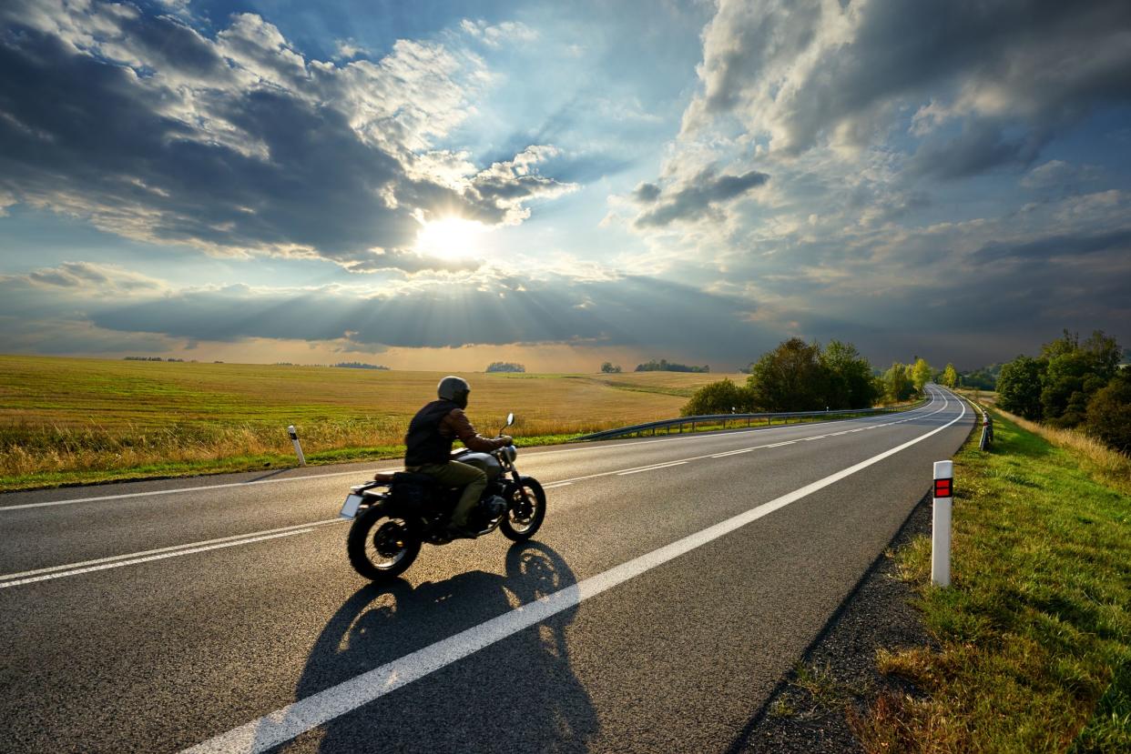 Man on motorcycle riding on asphalt road in rural landscape at sunset with dramatic clouds