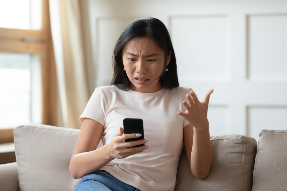 Annoyed Asian girl sit on couch in living room looking at her phone, illustrating a story on Singapore online home rental scams.