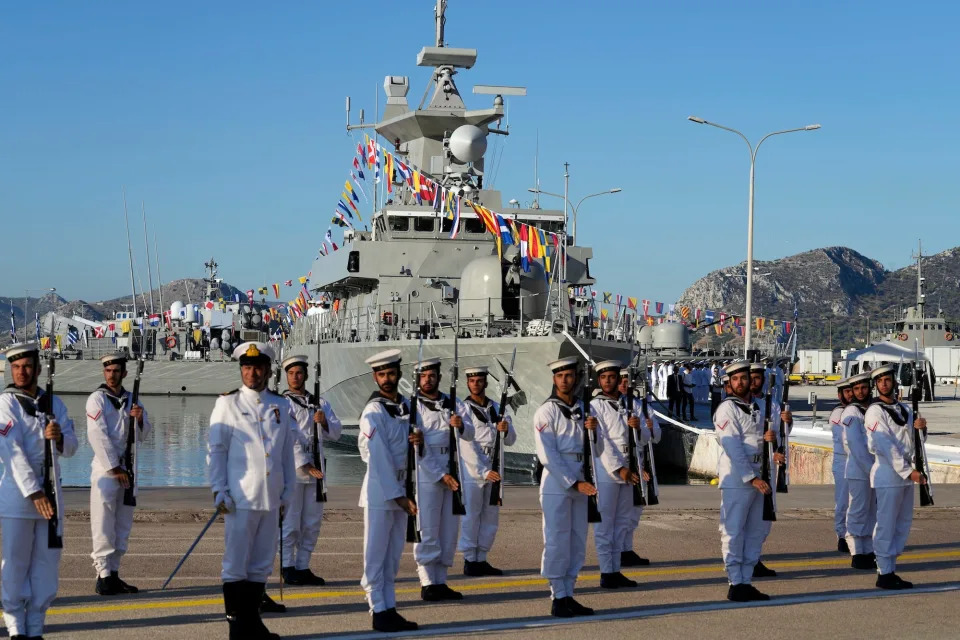 Greek soldiers in uniform stand in front of a warship with a blue sky