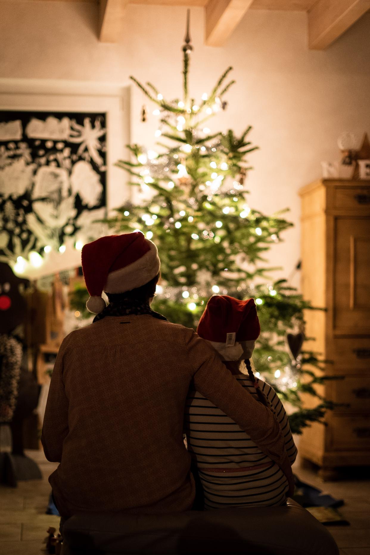  The silhouettes of a parent and young child both wearing Santa hats. They are out of focus, with a christmas tree lit in gold lights in focus in the background. 