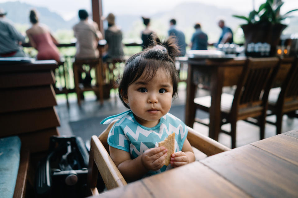 A young child with a patterned top sits in a high chair at an outdoor café, holding a piece of bread. People are seen in the background standing by a railing