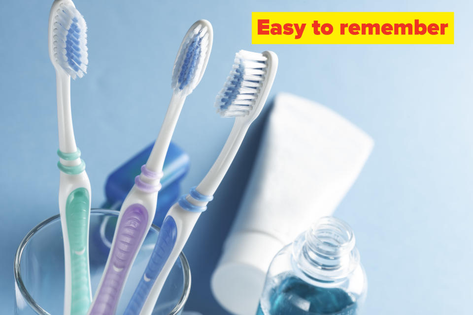 The top of three toothbrushes sticking out of the top of a clear glass holder pictured next to an opened bottle of mouthwash with a tube of toothpaste and dental floss container in the background