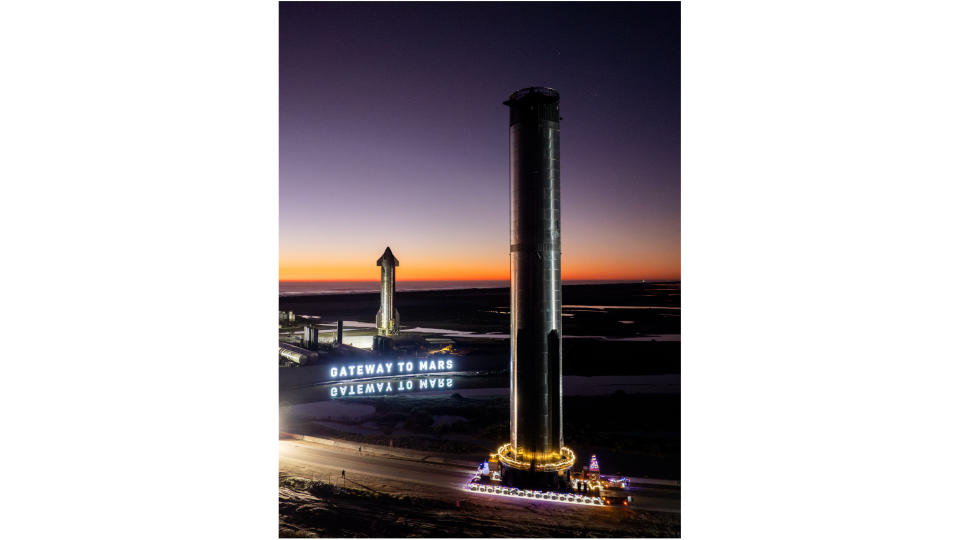 a rocket booster and upper stage are positioned vertically against a dusky sky, near a white light-up sign that says 