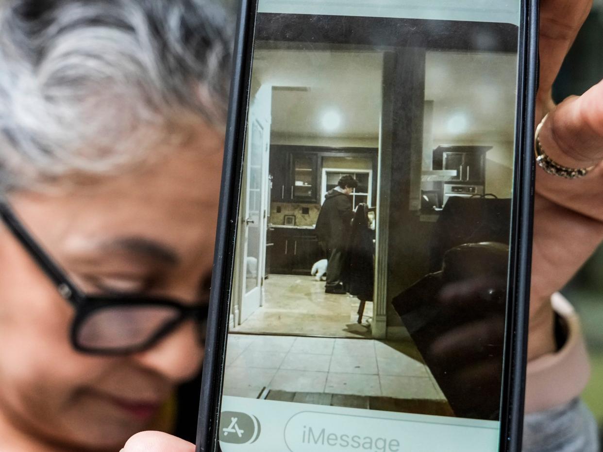 A woman with gray hair and glasses looks down and holds up a cellphone to the camera with a picture of a young man standing in a kitchen on it.