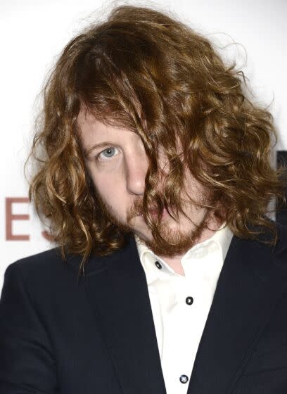 A man in a suit looks coyly out from behind his wavy red hair as it falls over his face