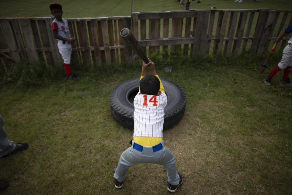 In this Aug. 12, 2019 photo, a young baseball player hits a tire with a bat during a practice at Las Brisas de Petare Sports Center, in Caracas, Venezuela. Boys run hard, lift weights, bat and pitch in the heat on the baseball field at next to the biggest slum in Caracas. (AP Photo/Ariana Cubillos)