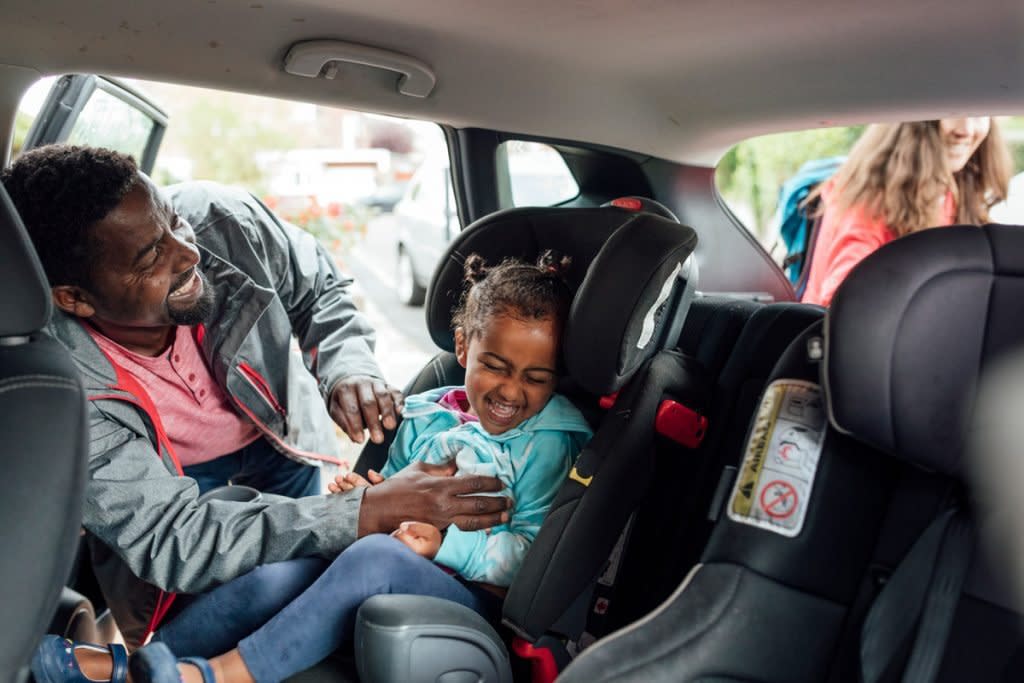 A father tickles is daughter as he puts her in her car seat with the mother putting things in the trunk. 