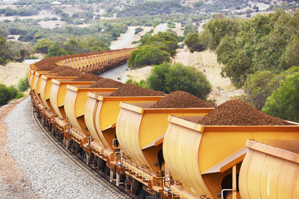 Heavy train loaded with brown hematite iron ore commodities in orange freight wagons passes through rolling hills.