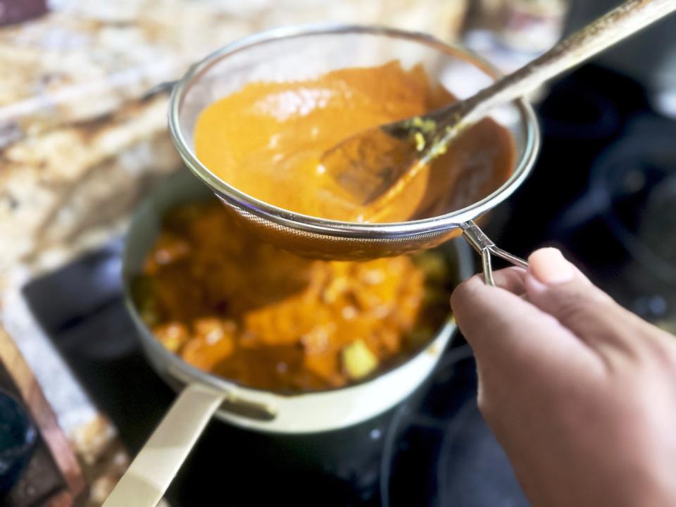 An orange sauce being strained through a sieve into a larger pan. A black stove range sits in the background