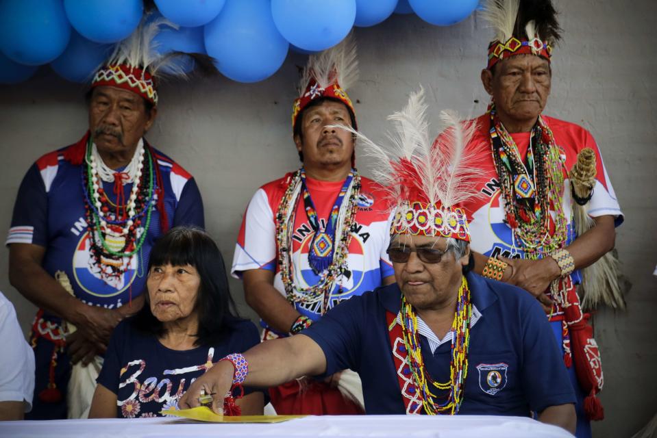 La líder indígena maká Tsiweyenki, cuyo nombre en español es Gloria Elizeche (delante, izquierda), asiste a una ceremonia con Mateo Martínez (derecha) en la que el gobierno entregó títulos de propiedad a familias maká, con hombres vestidos con trajes tradicionales escoltándolos, en Mariano Roque Alonso, Paraguay, el 22 de abril de 2019. Tras la muerte del esposo de Tsiweyenki, que era el jefe de los maká, su viuda se convirtió en la primera mujer que hereda el puesto. “A la viuda de Chemei se le consultan todos los asuntos; ella debe dar su aprobación o rechazo", explicó Martínez. (AP Foto/Jorge Saenz)