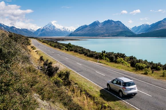 Car driving along a highway near a glacial lake with snow-capped mountains in the distance.