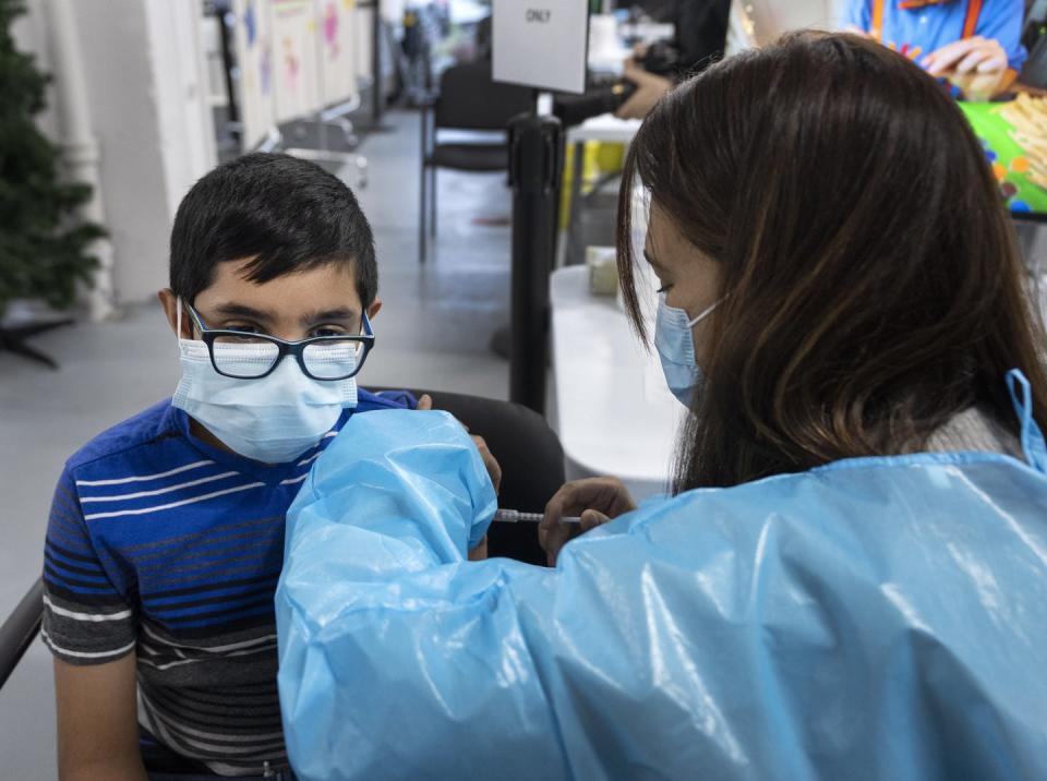 A boy wearing glasses and a mask gets vaccinated by a health-care worker wearing PPE