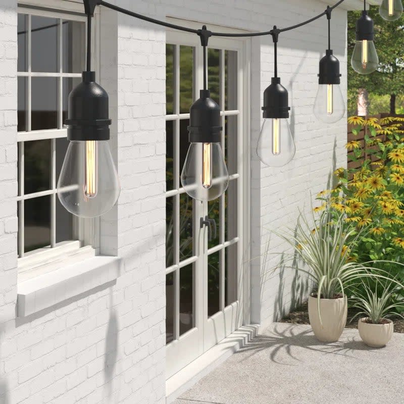 Outdoor string lights hanging above a patio beside a white brick wall with a window and plants