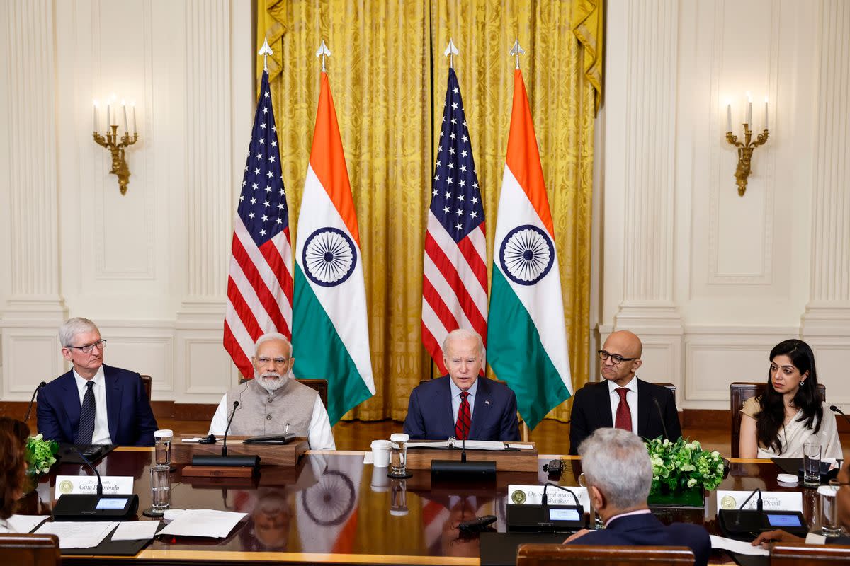 A white man sits in the middle of a table, surrounded by people on either side of him. In the background are two flags with red and white stripes and white stripes on a blue square, as well as two flags with orange, white and green stripes. On the white stripe of the flag is a spoked wheel.  Anna Moneymaker/Staff, Getty Images