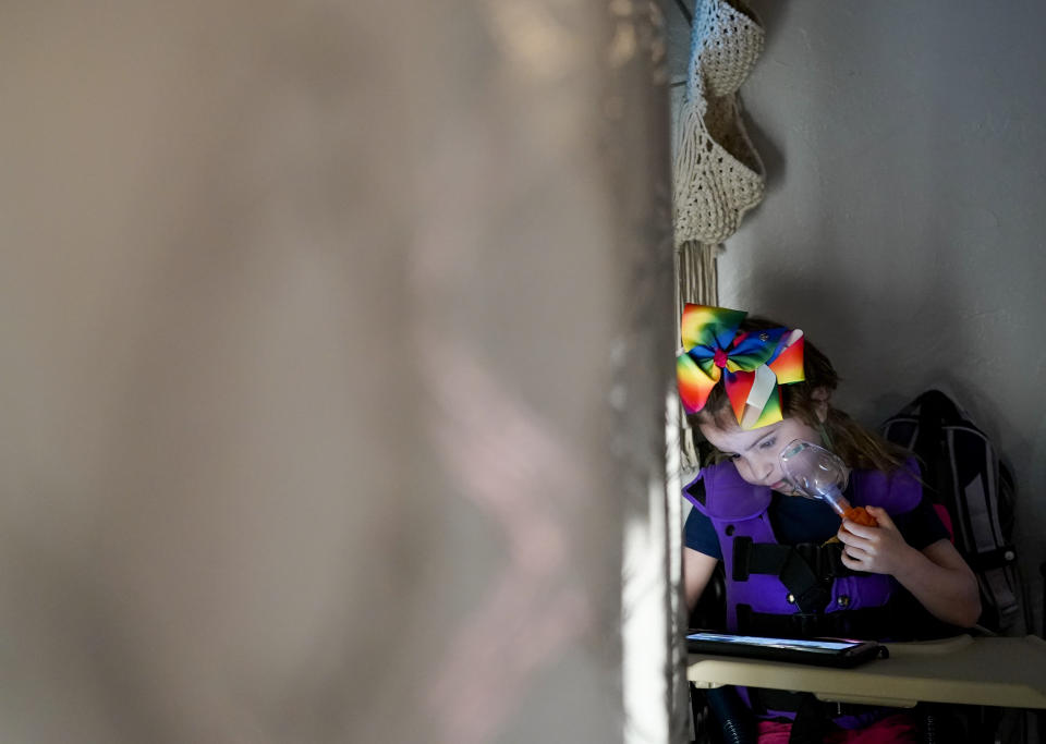 Scarlett Rasmussen, 8, watches a video on her tablet while using an airway clearance system that uses vibrations to clear mucus from her lungs while getting ready for school, Wednesday, May 17, 2023, at their home in Grants Pass, Ore. Scarlett's mother, Chelsea, says, “Every day I woke up with this immense burden: ... My child does not feel like she belongs. She can’t access a meaningful education. And there’s absolutely nothing I can do about it. ... I felt like I was drowning. ... We wasted a year with a child that could do grade-level work.” (AP Photo/Lindsey Wasson)