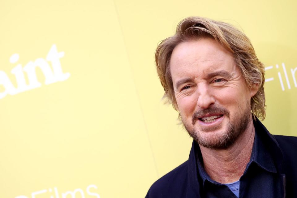 owen wilson smiling and looking off camera with a yellow wall in the background