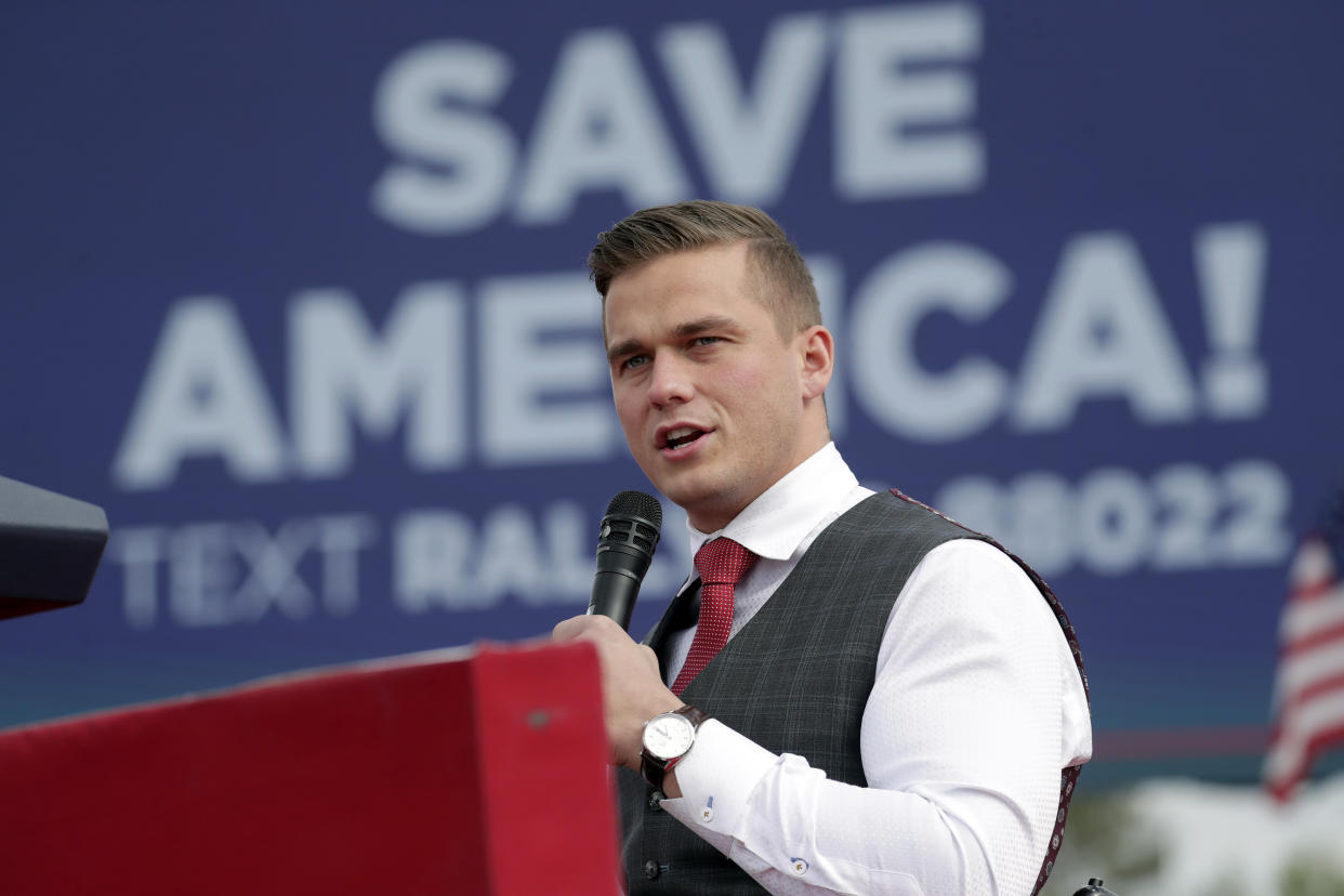 A neatly brushed and groomed Rep. Madison Cawthorn takes the microphone in front of a poster that says: Save America.