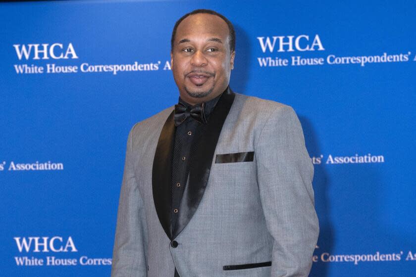 Roy Wood Jr. in a grey suit with a black suit shirt and tie posing with his hands at his side in front of a blue background.