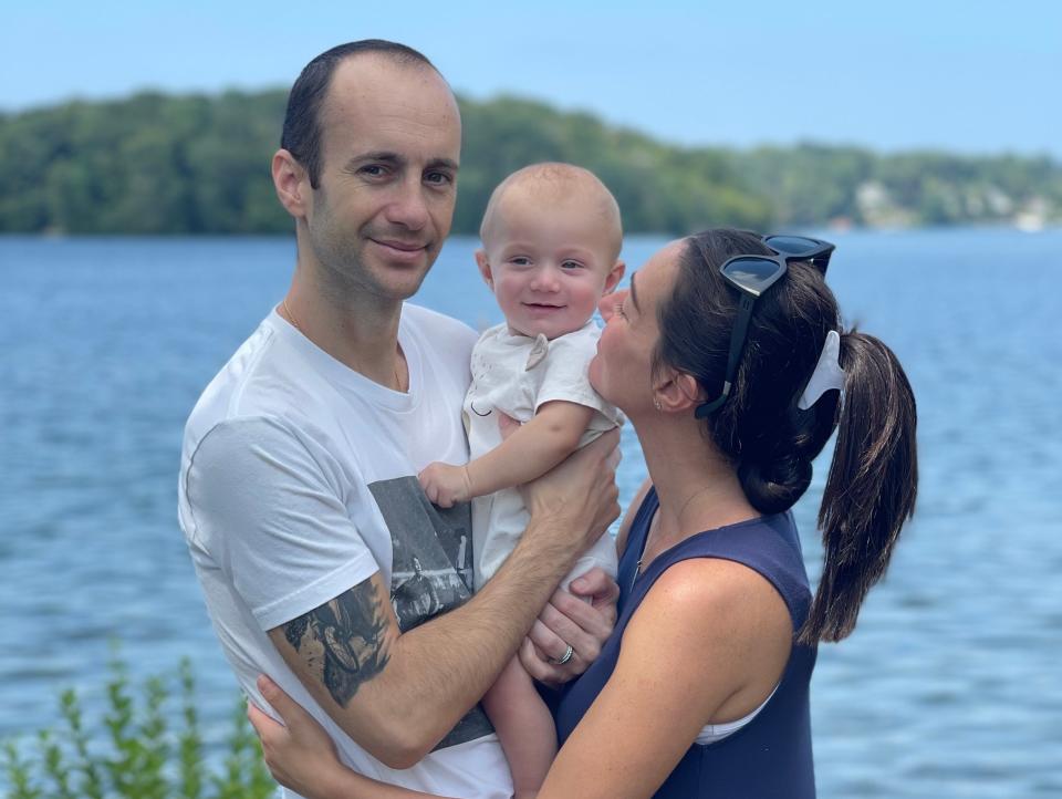 Anthony Di Laura and his wife, Jackie Cucullo, wrap their arms around each other and their first child, their baby son, JP, in front of a wide river.