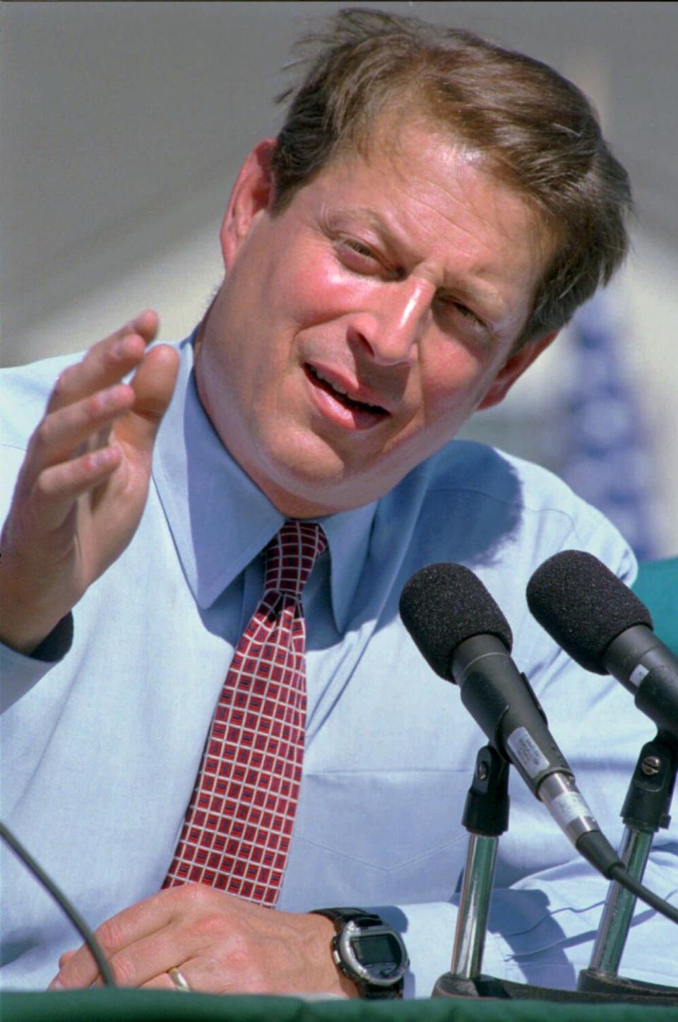 A vertical head-and-shoulders frame of Al Gore in front of microphones, gesturing with his right arm