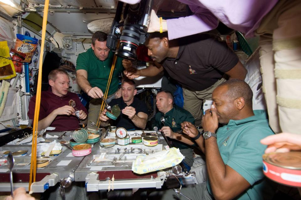 Several people gather around a table with cans and other food on it at the International Space Station